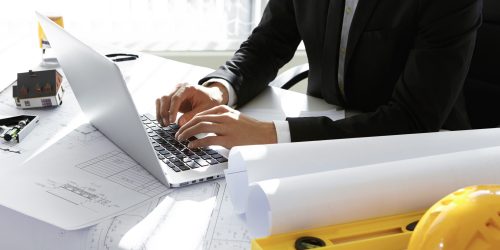 Cropped view of young Caucasian engineer wearing formal suit working on construction project using laptop computer, sitting at office desk, surrounded with engineering tools and rolls of blueprints